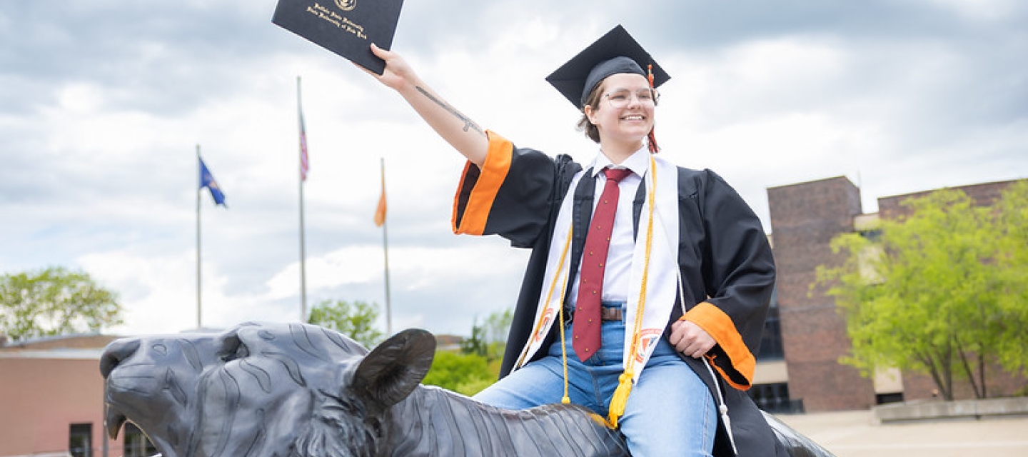 Student on the back of a tiger statue in cap and gown holding diploma