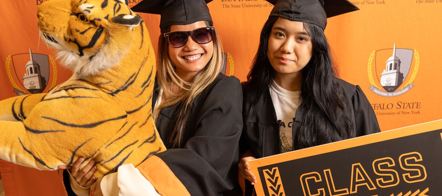 Two students in cap and gown holding a stuffed animal tiger